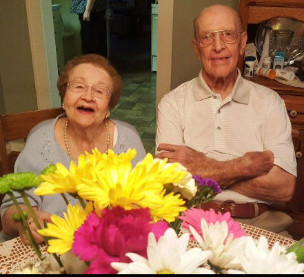a short woman sits on the right of a tall man both are in their late eighties and smiling. there are flowers in the foreground